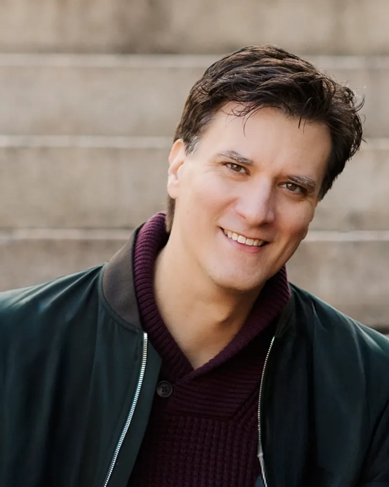 Axel Gimenez, smiling, in a dark green bomber jacket over a burgundy sweater, on a flight of stone steps.